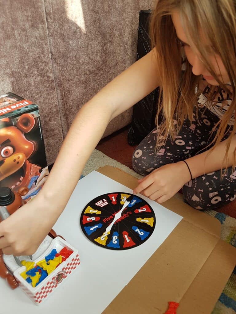 A young girl wearing patterned leggings plays a board game. She spins a colourful spinner with numbers and animal shapes. A game piece shaped like a bear with a tray of yellow and blue figures sits nearby; a "Five Nights at Freddy’s" game box is visible in the background.