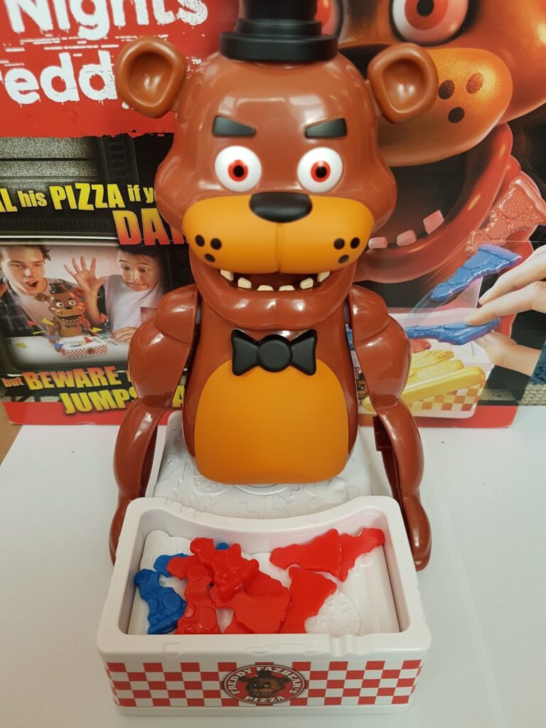 A plastic Five Nights at Freddy’s bear toy sits upright with a surprised expression. In front of it is a white tray containing red and blue plastic pizza slice pieces. The background shows the game’s packaging with images and bold text.
