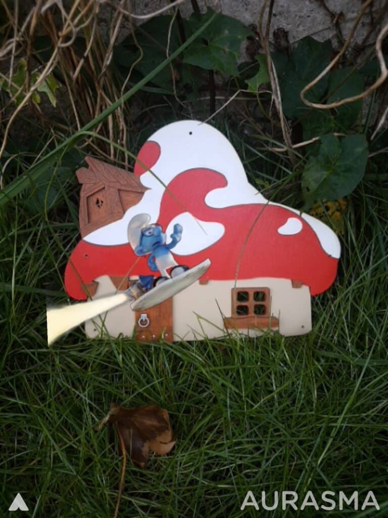 A small Smurf figurine rides a yellow surfboard in front of a whimsical red-and-white mushroom house model, set on green grass with leaves and vines in the background. The word “AURASMA” appears in white text at the bottom right.