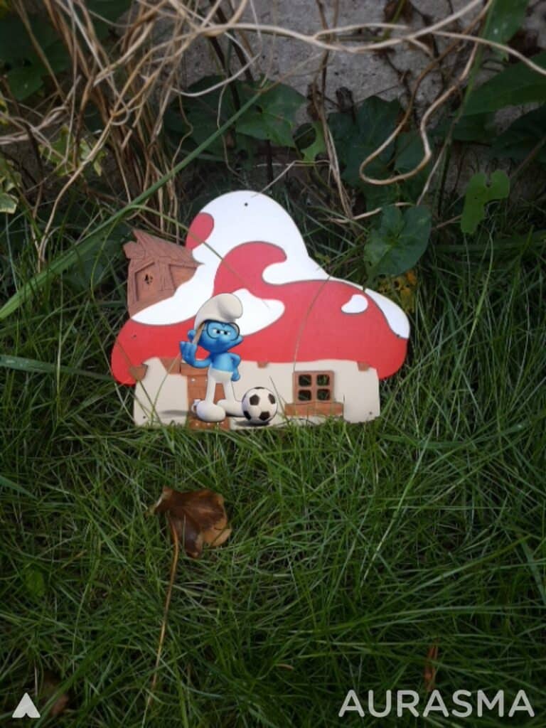 A small Smurf figurine with a red rucksack and a football stands in front of a decorative mushroom house in green grass, surrounded by leafy plants and vines. The image has the word "AURASMA" in white text in the bottom right corner.
