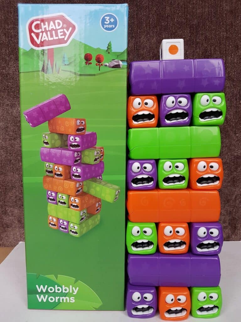 A box labelled “Chad Valley Wobbly Worms” stands beside a stacked tower of colourful blocks. The blocks are purple, green, and orange with cartoon worm faces showing various surprised and happy expressions. The game is for ages 3+.