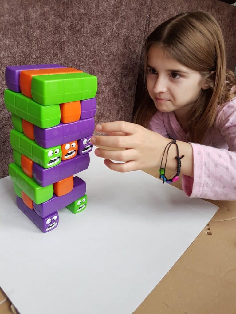 A young girl with long brown hair and a pink top concentrates as she plays a stacking game with colourful purple and green blocks, each displaying cartoon faces with various expressions, on a white surface against a brown background.