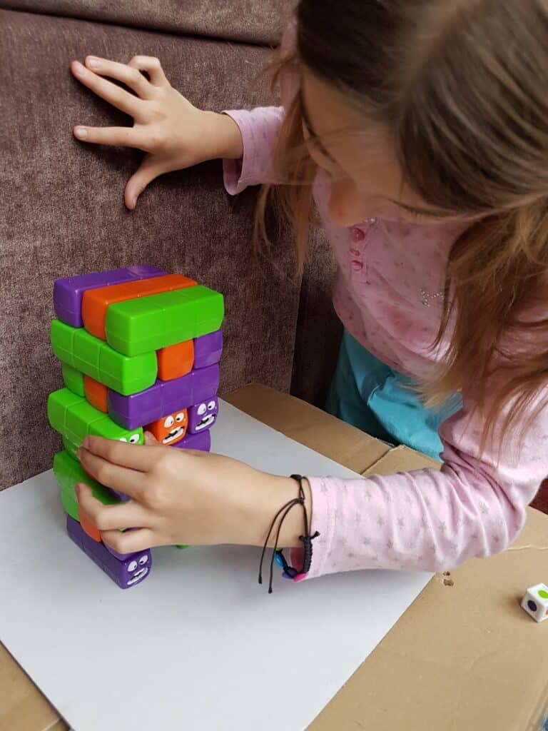 A child with light brown hair and a pink shirt carefully removes a green block from a colourful, stacked block game. The blocks are red, green, purple, and have cartoon faces. The game is set up on a piece of white paper on a cardboard surface.
