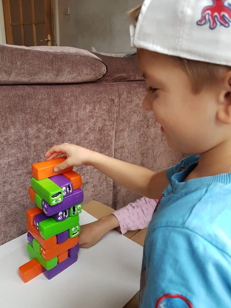 A young boy in a blue shirt and white cap smiles whilst playing a stacking game with colourful blocks featuring cartoon faces. Another child’s arm, wearing a pink sleeve, helps steady the stack. They are next to a brown sofa indoors.