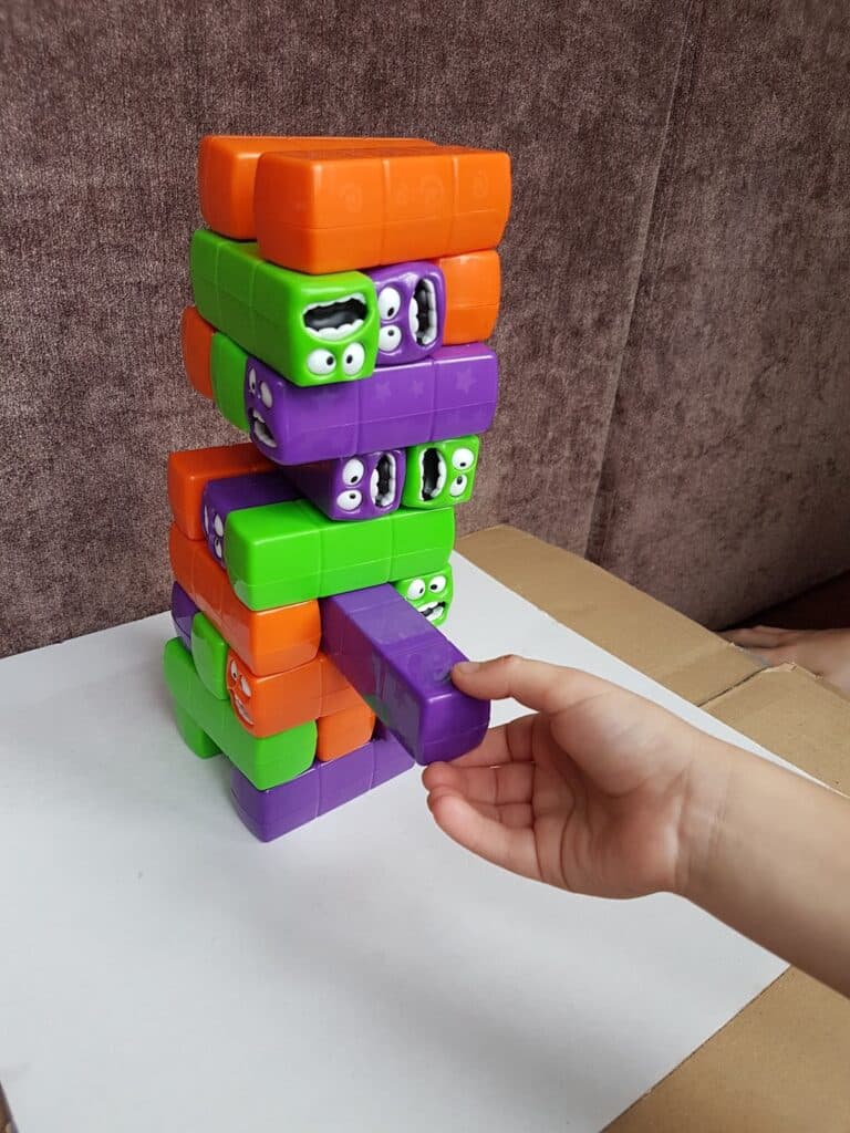 A child’s hand pulls a purple block from a colourful Jenga-like tower made of green, orange, and purple blocks, each featuring cartoon faces. The game rests on a white surface with a brown background.