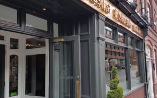 The photo shows the exterior of a restaurant called Cucina Rustica, with large glass windows, a dark grey entrance, and gold signage above the door. A spiral-shaped topiary in a tall planter stands beside the entrance on a brick pavement.