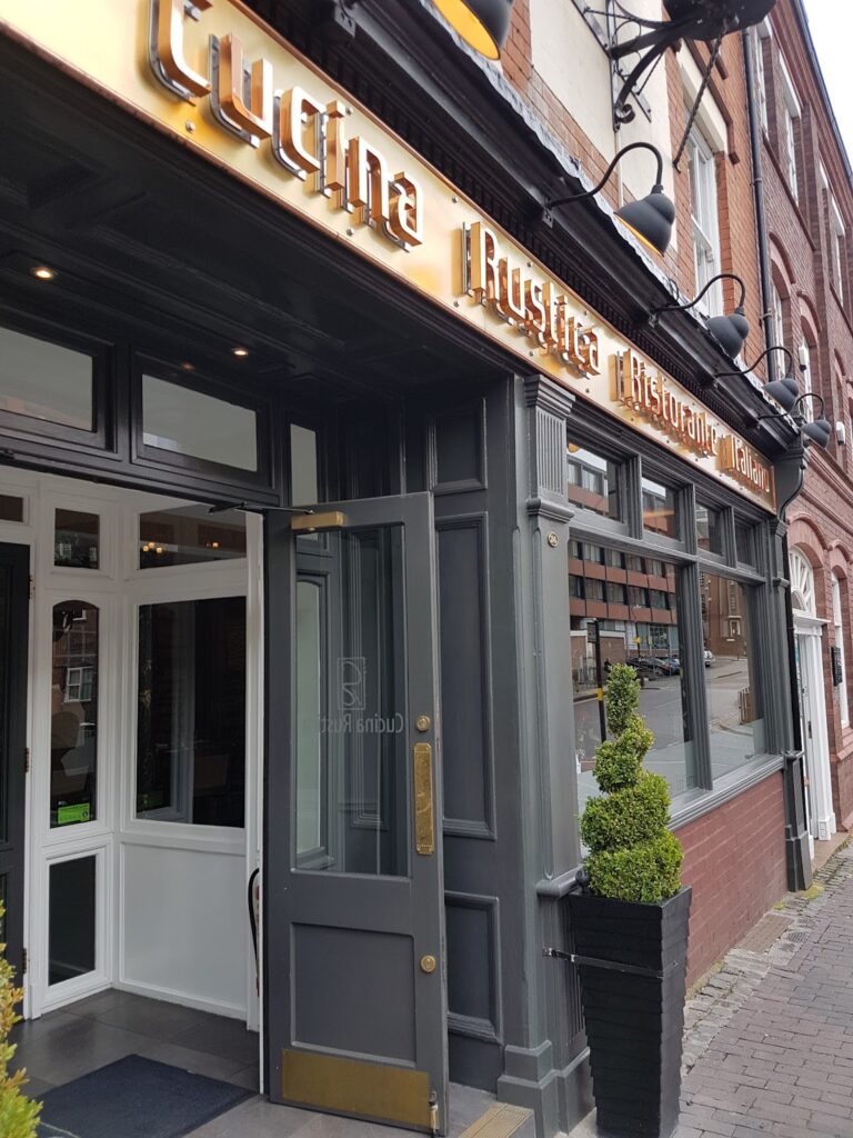 The image shows the exterior of a restaurant named “Cucina Rustica”, with gold signage above a dark grey facade. The front door is open, revealing part of the interior, and a topiary plant in a tall pot stands by the entrance. Brick buildings line the street outside.
