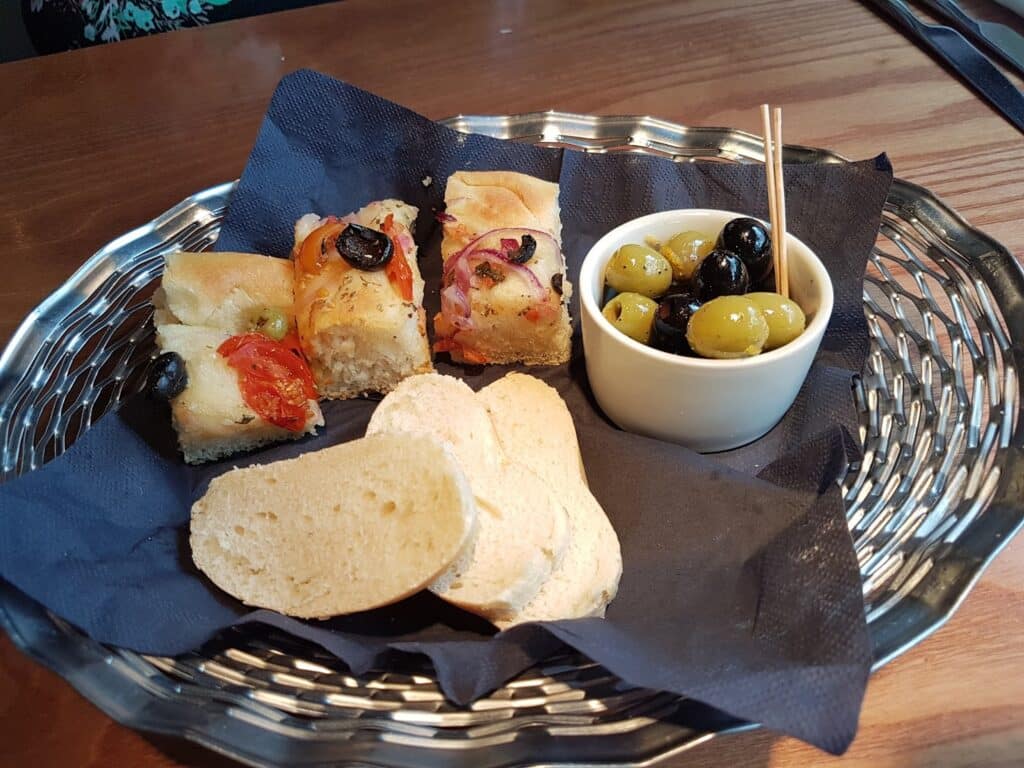 A silver basket holds slices of white bread, three pieces of focaccia topped with tomatoes and olives, and a small cup filled with green and black olives, all resting on a dark blue serviette on a wooden table.