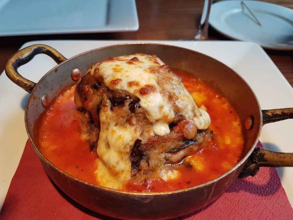 A serving of baked lasagne topped with melted, browned cheese sits in a rustic copper pan filled with tomato sauce. The dish is placed on a red serviette on a white plate, with an empty plate and cutlery in the background.