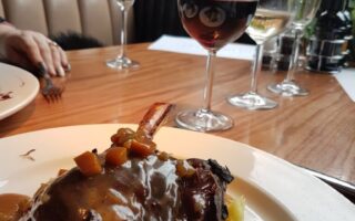 A plate with braised meat in brown gravy, garnished with diced vegetables, sits on mashed potato. Behind it are three glasses—one of red wine, one of white wine, and one empty—on a wooden table by a window with city buildings visible outside.