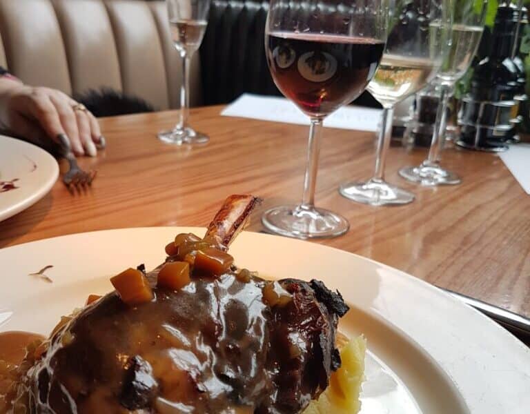 A plate with braised meat in brown gravy, garnished with diced vegetables, sits on mashed potato. Behind it are three glasses—one of red wine, one of white wine, and one empty—on a wooden table by a window with city buildings visible outside.