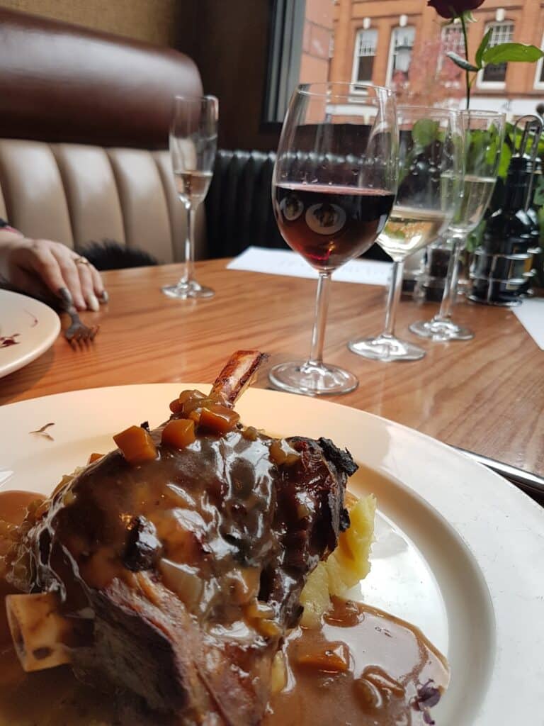A close-up of a plated dish with a bone-in meat cut topped with gravy and diced vegetables, served on mashed potato. In the background are wine glasses with red and white wine, a champagne flute, and a hand resting on the table near a window with a city view.