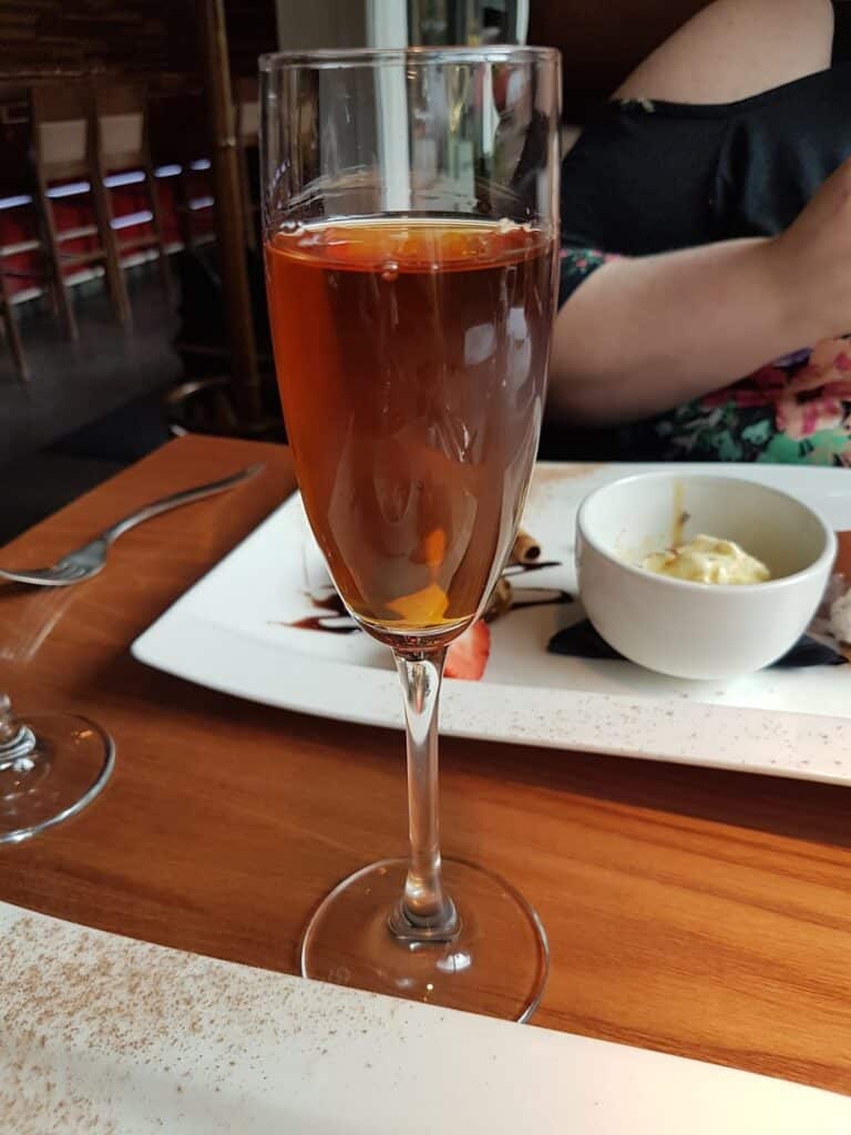 A tall champagne flute filled with a reddish-brown sparkling drink, garnished with an orange twist, sits on a wooden table set for dining. Behind it, there is a rectangular white plate with pudding and sauce, and a person in a floral top.
