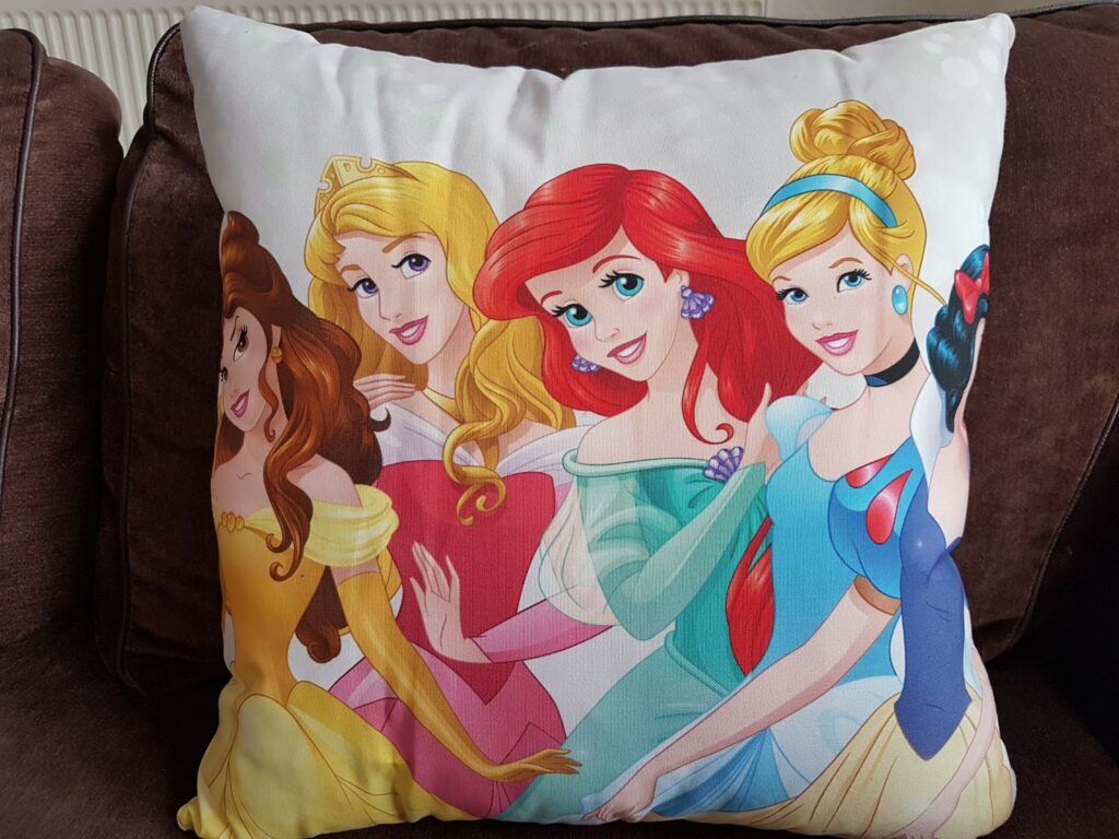 A square cushion with illustrations of four Disney princesses—Belle, Aurora, Ariel, and Cinderella—smiling and posing together on a white background, resting on a brown sofa. The colourful designs feature their signature dresses and hairstyles.