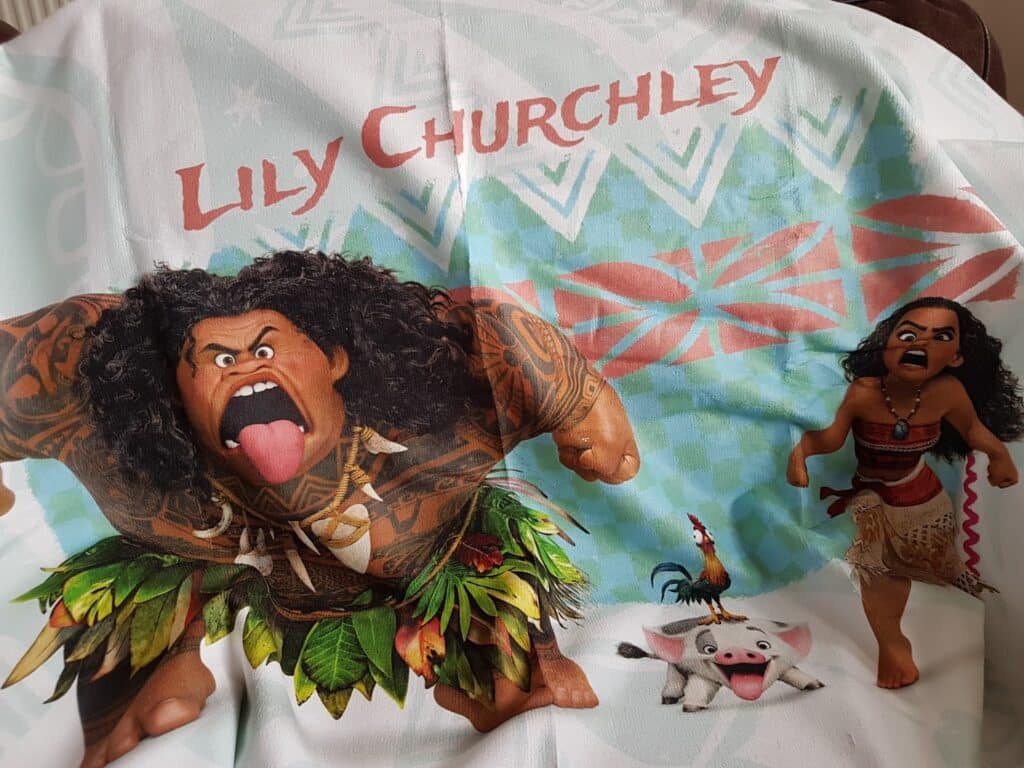 A blanket featuring characters from Disney’s Moana. Maui is flexing and sticking out his tongue on the left, whilst Moana, also with her tongue out, stands on the right. Hei Hei the cockerel and Pua the pig are in the centre. The name "Lily Churchley" appears at the top.