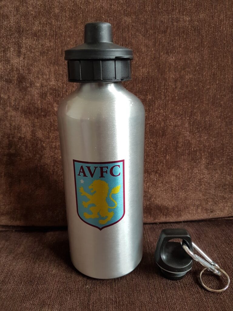 A silver water bottle with the Aston Villa football club crest, showing a yellow lion and "AVFC" text, sits on a brown fabric surface. Next to it is a detached black lid with a metal keyring.