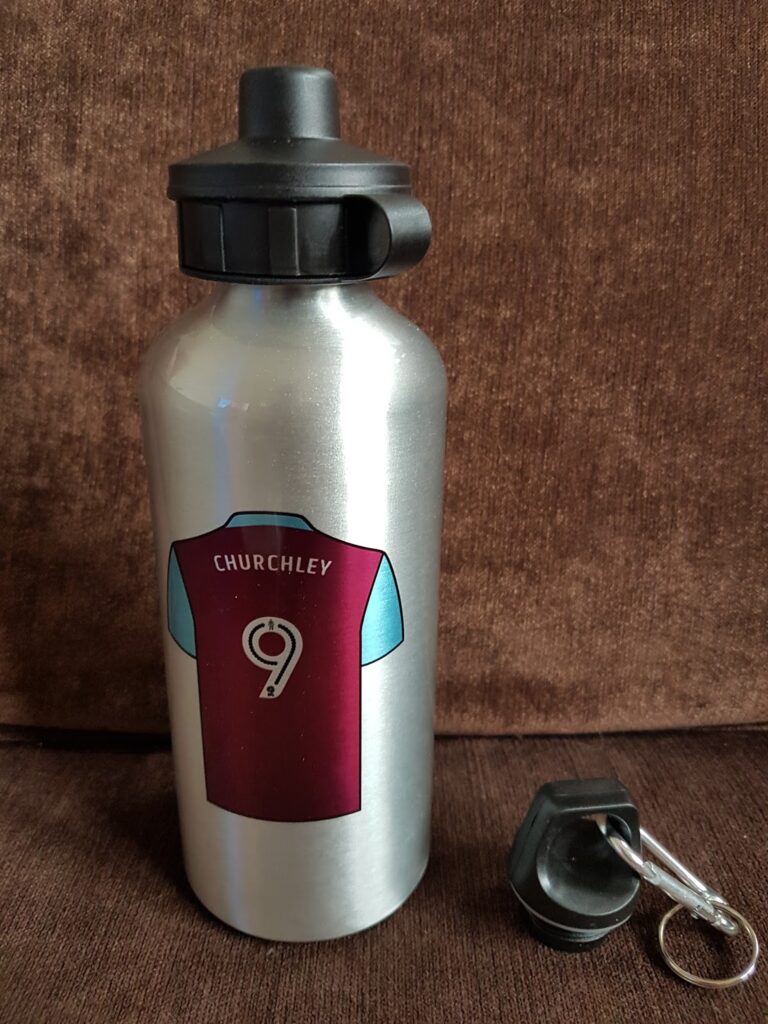 A silver metal water bottle with a maroon and blue football shirt graphic. The shirt displays the name "Churchley" and number 9. The bottle cap is black; a second black cap with a keyring attachment sits beside the bottle on a brown fabric surface.