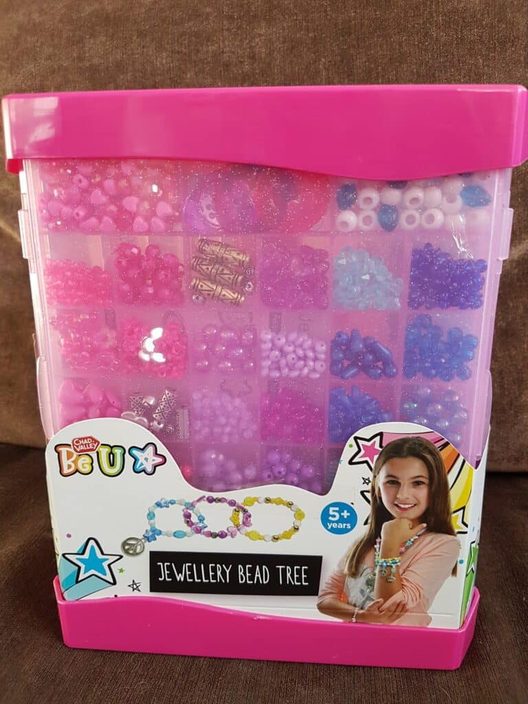 A plastic container with a pink lid holds assorted jewellery beads in pink, purple, and blue tones. The packaging features a smiling girl wearing bead bracelets and a sign reading "Jewellery Bead Tree, 5+ years." The background is brown fabric.