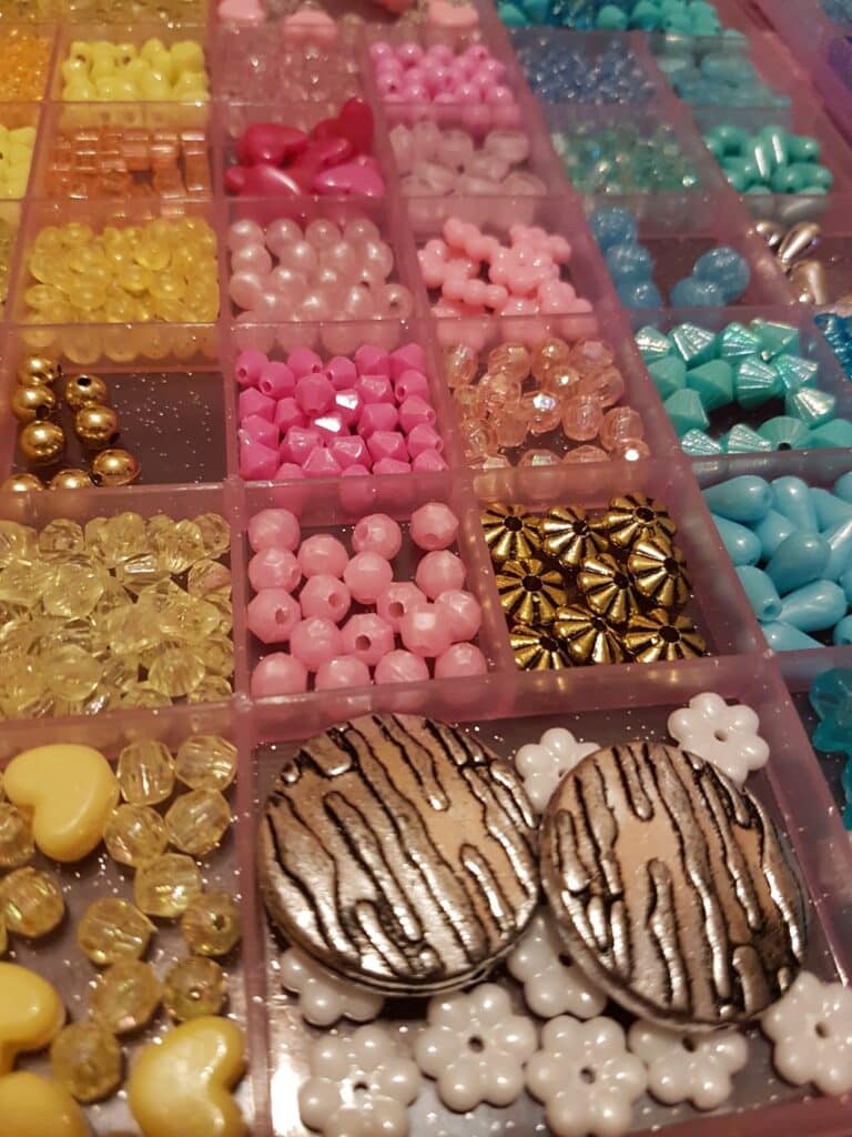 A plastic organiser box with small compartments holds various colourful beads and charms, including round, heart, flower, and star shapes in pink, yellow, blue, and gold. Two large, round beads with a gold and black striped pattern are in one section.