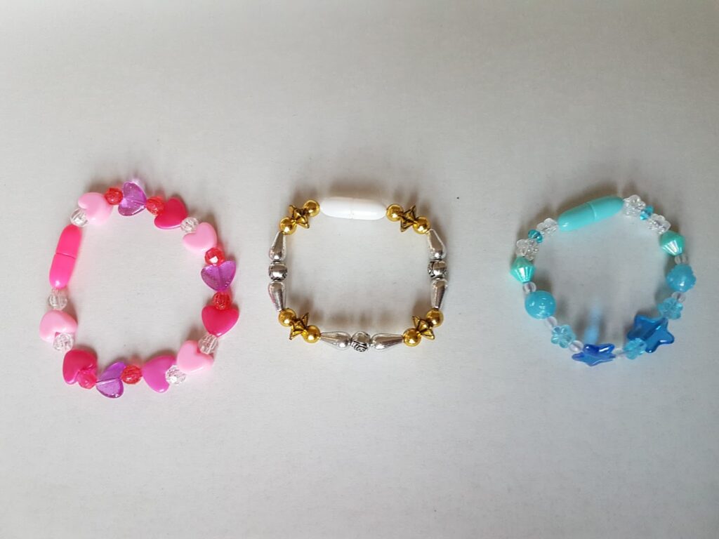 Three beaded bracelets on a white surface: the left one is pink with heart-shaped and round beads, the middle one is gold and silver with a white oval bead in the centre, and the right one is blue with butterfly and round beads.