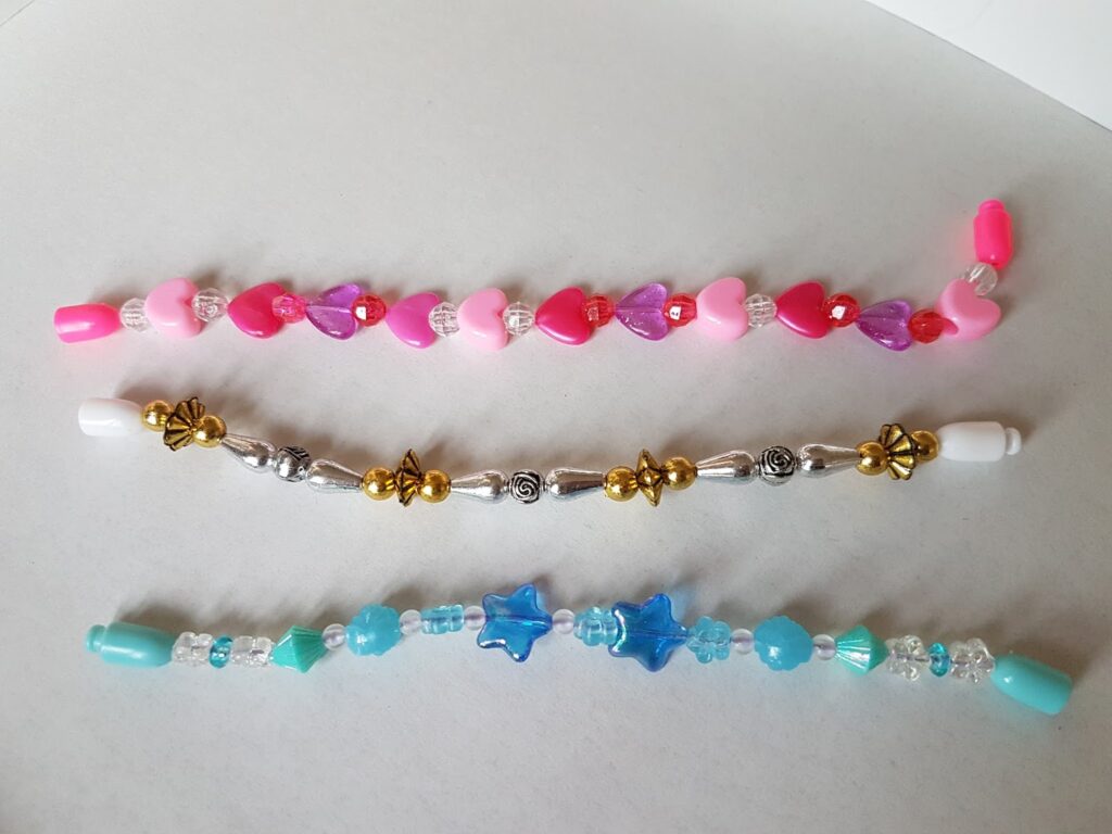 Three colourful beaded bracelets are laid out on a white surface. The top bracelet has pink, red, and purple heart beads; the middle has gold and silver beads with flower designs; the bottom features blue star beads and other blue and clear beads.