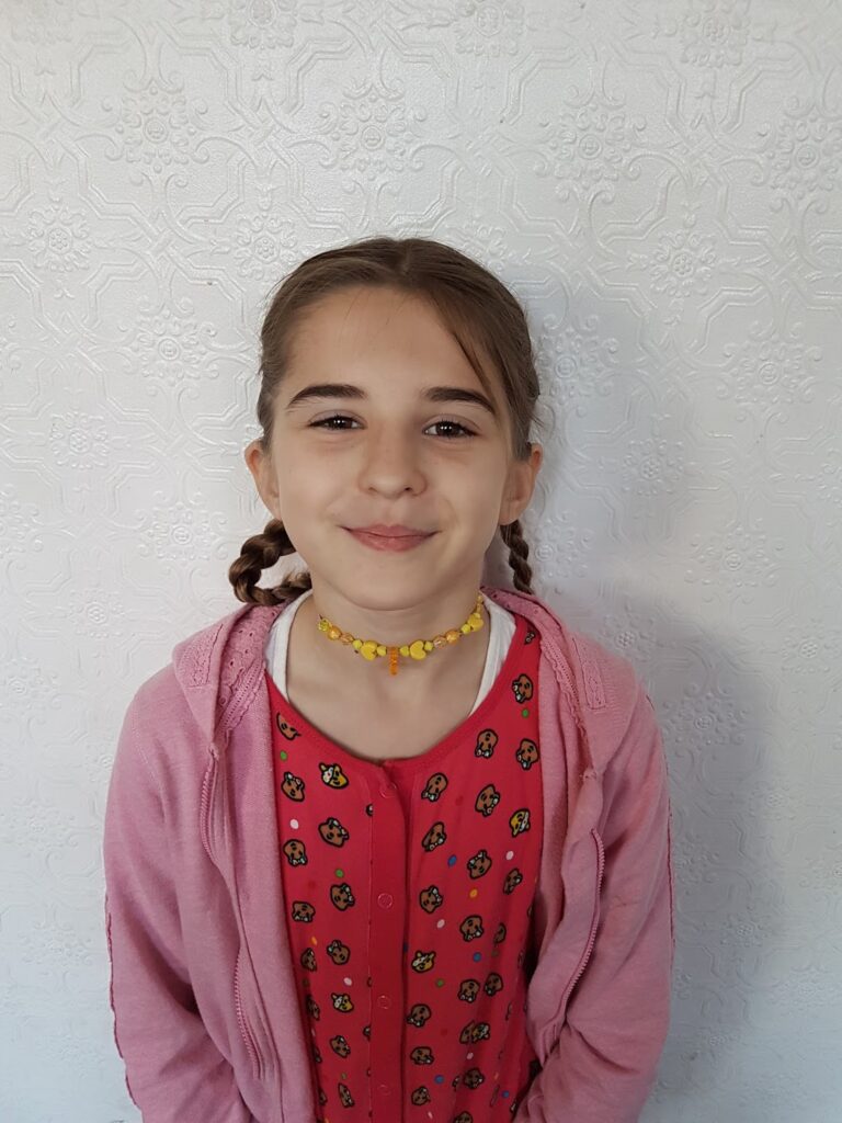 A young girl with fair skin and brown hair in plaits stands against a textured white wall. She wears a pink hoodie over a red shirt with colourful cartoon patterns and a yellow beaded choker. She smiles softly, looking at the camera.