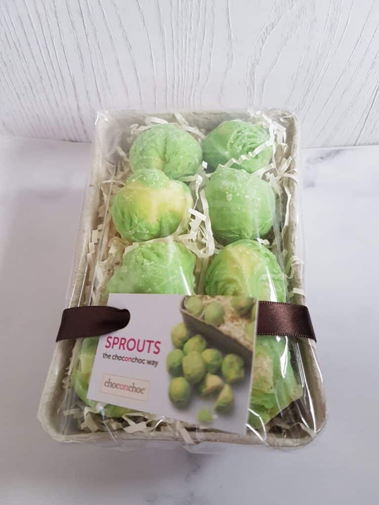A cardboard tray holds six green chocolates shaped like Brussels sprouts, nestled in shredded paper. A brown ribbon secures a label reading “SPROUTS the choconchoc way” with a photo of chocolate sprouts and the Choconchoc logo.