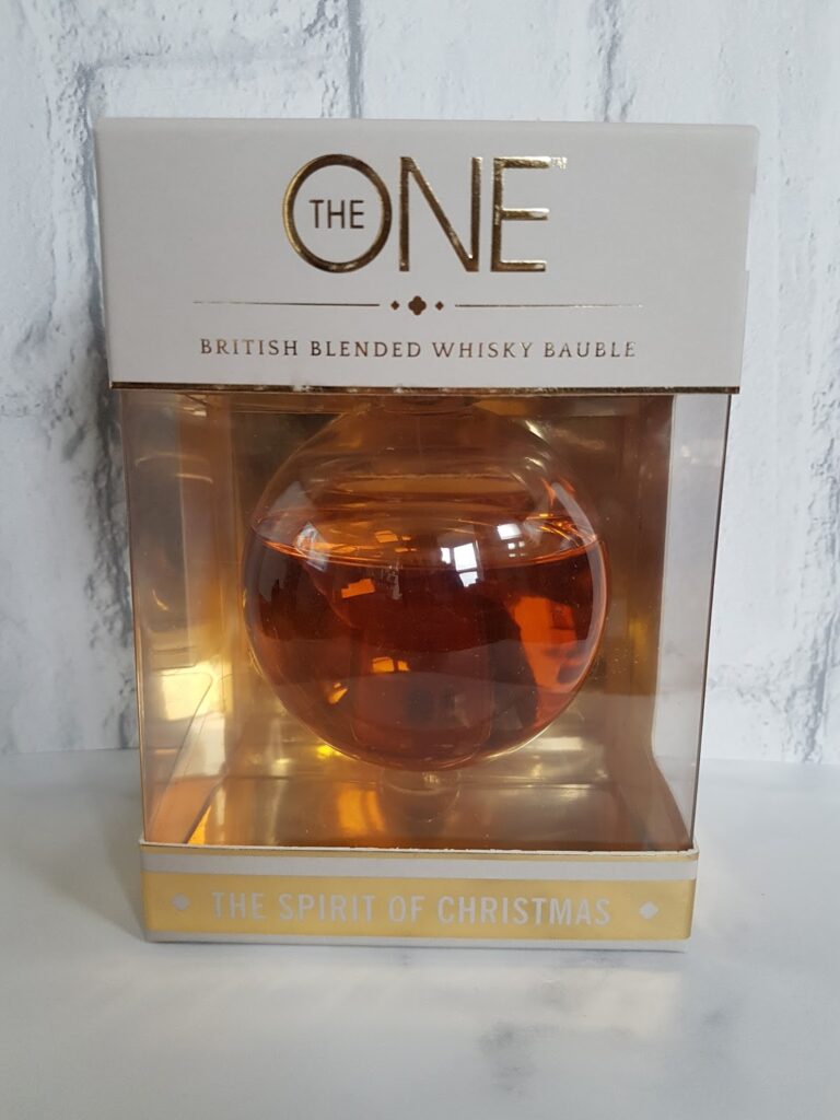 A round glass bauble filled with amber whisky is displayed in a transparent box. The packaging reads "THE ONE British Blended Whisky Bauble" in gold lettering and "The Spirit of Christmas" at the bottom. The background is a white textured wall.