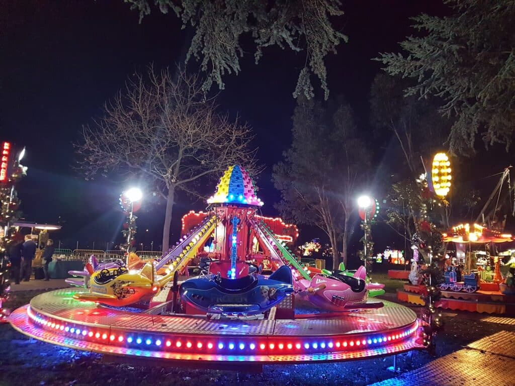 A brightly lit children's funfair ride with colourful seats shaped like animals spins under night trees. The ride is surrounded by vibrant lights, and other illuminated attractions are visible in the background. People stand nearby under the night sky.