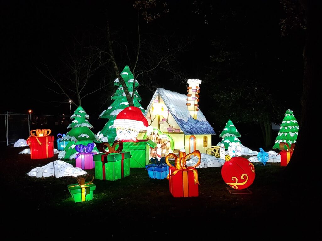 A festive night-time scene with a glowing cottage surrounded by snow, large decorated Christmas trees, and oversized, colourful presents with bows. A giant Father Christmas hat sits on the cottage, creating a magical holiday atmosphere.
