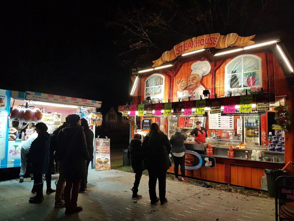 A nighttime outdoor market scene with people standing at two brightly lit food stalls. The right stall, “STEAKHOUSE,” has a chef illustration and festive decor. The left stall sells sweets. Trees and darkness are visible in the background.