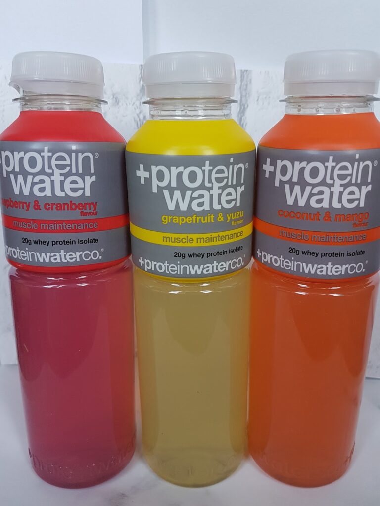 Three clear bottles of +Protein Water stand side by side. The flavours are raspberry & cranberry (pink drink), grapefruit & yuzu (yellow drink), and coconut & mango (orange drink). Each bottle highlights 20g whey protein isolate for muscle maintenance.