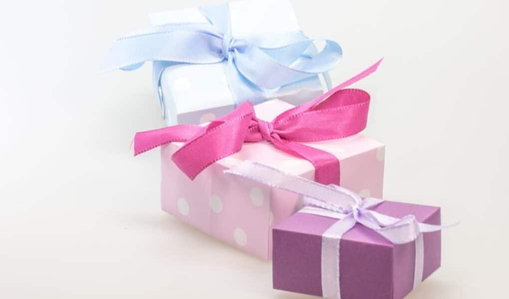 Three gift boxes are stacked together. The top box is wrapped in white paper with a light blue ribbon, the middle box is pink with white polka dots and a bright pink ribbon, and the bottom box is purple with a white ribbon. All are set against a plain background.