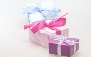 Three gift boxes are stacked together. The top box is wrapped in white paper with a light blue ribbon, the middle box is pink with white polka dots and a bright pink ribbon, and the bottom box is purple with a white ribbon. All are set against a plain background.