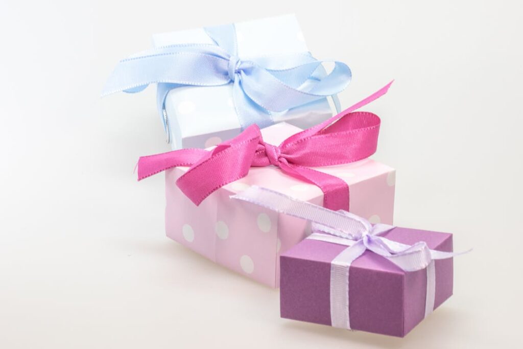 Three gift boxes are stacked on a white surface. The top box is light blue with white polka dots and a blue ribbon, the middle is pink with white polka dots and a pink ribbon, and the bottom is purple with a light purple ribbon.