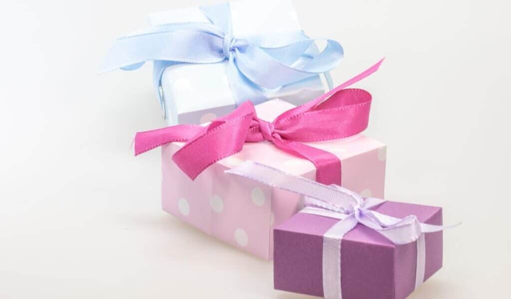 Three gift boxes are stacked closely together. The top box is light blue with a matching ribbon, the middle box is pink with white spots and a bright pink ribbon, and the bottom box is purple with a white ribbon, all against a plain white background.