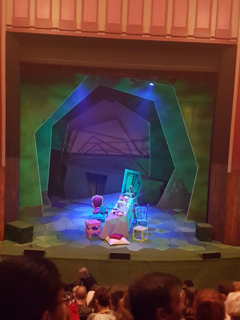 A stage set for a play features a whimsical scene with a long table, teacups, chairs, and colourful props. The backdrop is painted in shades of green, with geometric shapes suggesting a magical or fantastical setting. Audience members sit in the foreground.