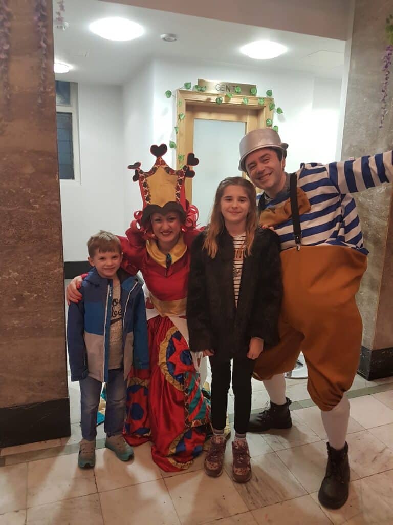 Two children stand smiling with two adults in colourful costumes. One adult is dressed as the Queen of Hearts, wearing a red and gold dress and a crown. The other wears a sailor outfit with striped shirt and braces. They are indoors, in front of a door labelled “GENTS.”.