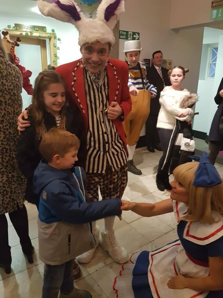 Two children, a girl and a boy, greet a woman dressed as Alice from "Alice in Wonderland," who is kneeling and shaking the boy’s hand. Behind them stands a smiling man in a red coat and bunny ears, with other people and performers in the background.