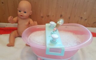 A naked baby doll sits on a white towel next to a pink and blue toy bath filled with soapy bubbles. The bath has toy taps and a silver shower head. The background is wood panelling.