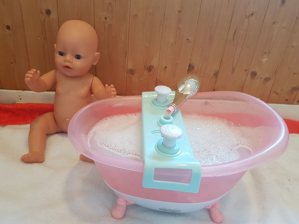 A plastic baby doll sits upright on a white towel with a red edge, next to a small pink toy bath filled with soapy water and bubbles. The bath has pretend tap handles and a toy shower head, set against a wooden wall background.