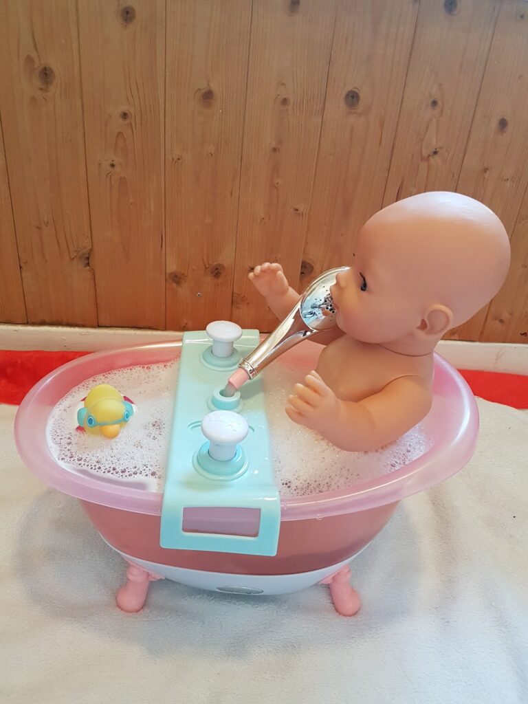 A bald baby doll sits in a pink toy bath filled with bubbly water, holding a silver shower head to its mouth. A small yellow rubber duck floats on the bubbles. The background is a wooden wall and a red-and-white towel is underneath the bath.