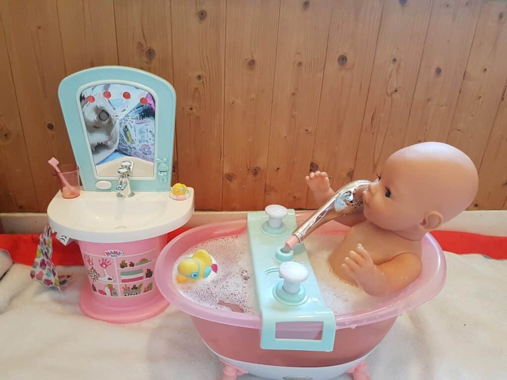 A baby doll sits in a pink and blue toy bath filled with bubbles, holding a shower head. Next to the bath is a toy sink with a mirror, a cup, and a towel. A yellow rubber duck floats in the bath. The background is wooden panelling.