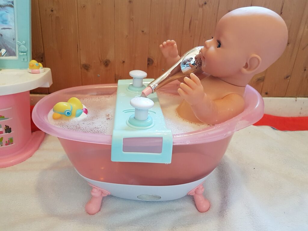 A plastic baby doll sits in a pink toy bath filled with bubbles, holding a toy hairdryer to its mouth. A yellow rubber duck floats in the bath, and the scene is set against a wooden wall with other toys nearby.
