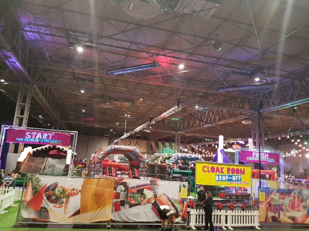 Indoor amusement event with colourful inflatable obstacle course under a high ceiling lit by fairy lights. Signs read "Start," "Finish," and "Cloakroom Drop-Off." White fences and printed banners enclose the area; some people are visible near the entrance.