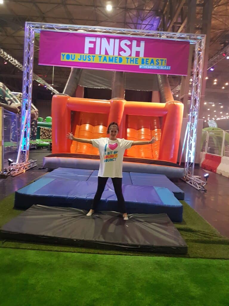 A smiling person stands with arms wide on mats in front of a large inflatable obstacle. Above them, a banner reads, "FINISH. You just tamed the beast!" The area is indoors with bright lights and green artificial grass. The person wears a colourful event t-shirt.