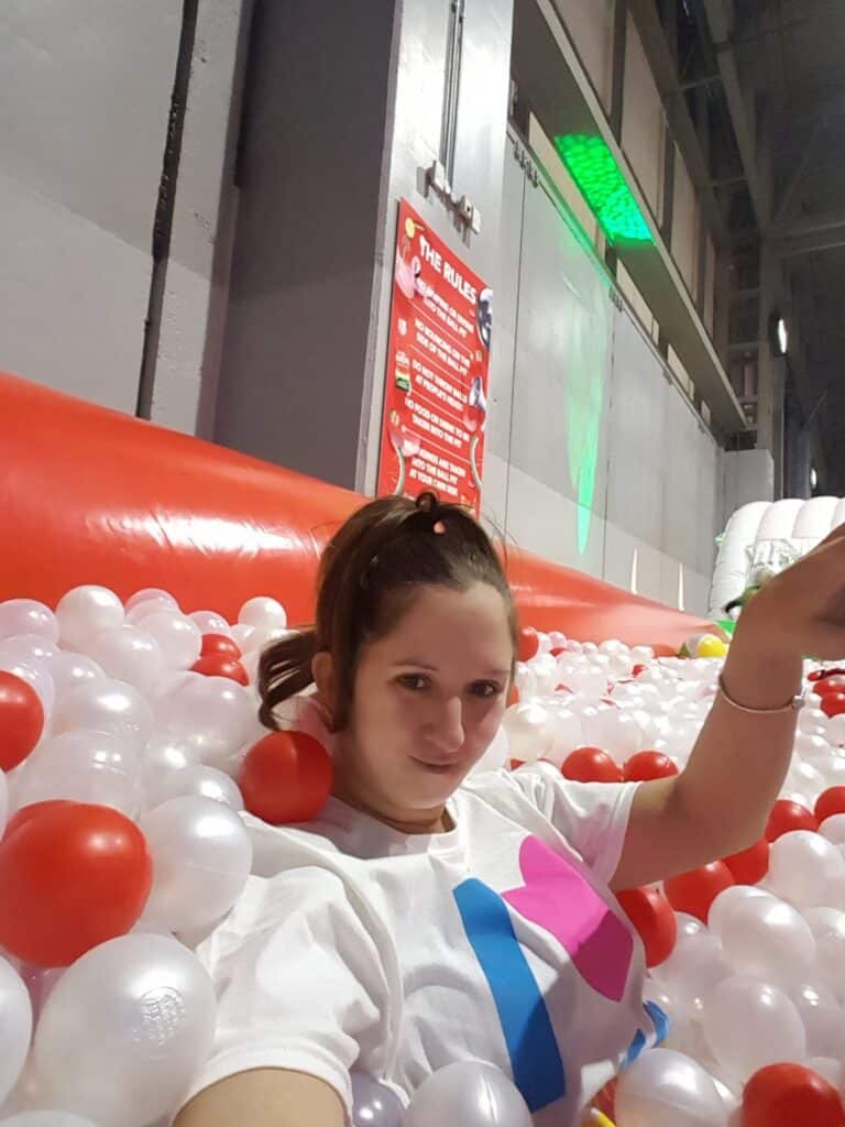 A woman with a ponytail takes a selfie while sitting in a ball pit filled with white and red balls. She wears a white shirt with colourful shapes. Behind her is a red inflatable barrier and a rules sign on a grey wall in an indoor play area.