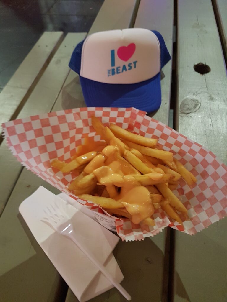 A tray of chips topped with melted cheese sits on a wooden picnic table. Next to the chips is a white paper container and fork. In the background, there is a blue and white lorry driver's hat with the phrase “I ♥ THE BEAST” printed on it.