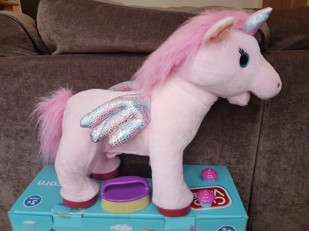 A plush pink unicorn toy with a shiny silver horn, pink mane and tail, and iridescent wings stands on a blue box. Two pink toy accessories and a yellow brush are next to it. The background is a brown sofa.