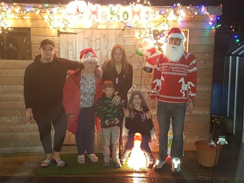 Six people stand outside a wooden shed decorated with Christmas lights. Two wear Father Christmas hats; one adult wears a Father Christmas beard and jumper. Two children are in front, one kneeling. A glowing sign and a Father Christmas decoration hang above, creating a festive atmosphere.